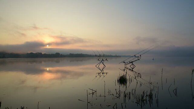 Carp fishing rods on rod pod at sunrise on misty lake, calm water reflections, summer dawn landscape in northern Italy