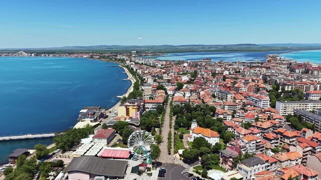 A city along the coast with many buildings and a clear blue sea. There are roads, trees, and a ferris wheel nearby. The day is sunny with few clouds.