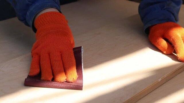 An unrecognizable person in blue work clothes and protective gloves is working on the processing of a wooden board. The process of sanding the surface of a board using sandpaper. Handicraft, hobby