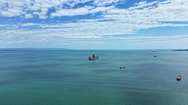 An industrial dredging vessel is working in open water. Nearby cargo ships are present. The sky is blue with scattered clouds above the scene.