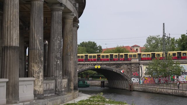 A red and yellow Berlin S-Bahn crosses a stone bridge over the water beside historic columns. Graffiti walls, trees and the riverside add urban contrast.