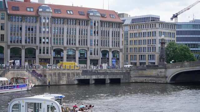 The Spree waterfront shows university buildings, a tour boat and a stone bridge in Berlin. Pedestrians, trees and rippling water create a lively city panorama.