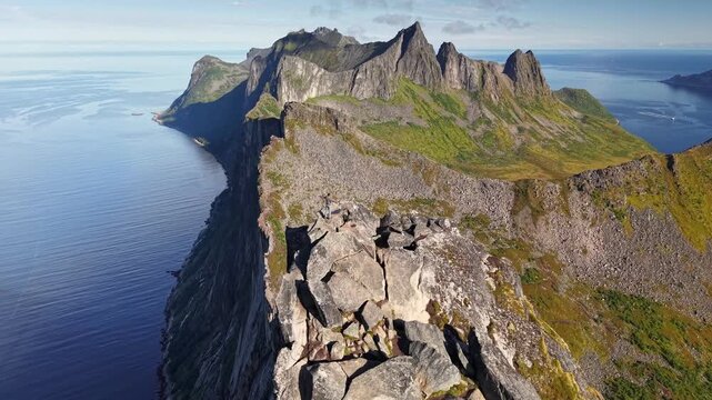 Brave tourist reaches sharp dangerous summit of Segla mountain in Senja, Norway. Huge steep ridge drops into vast blue ocean on Norwegian coast. Aerial 4K of extreme hiking adventure in wild Senja