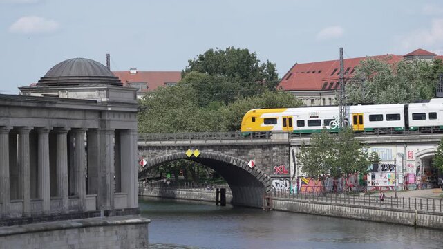 A green and white ODEG regional train crosses a stone canal bridge near a Berlin colonnade. Graffiti, water, trees and historic architecture add layered urban detail.