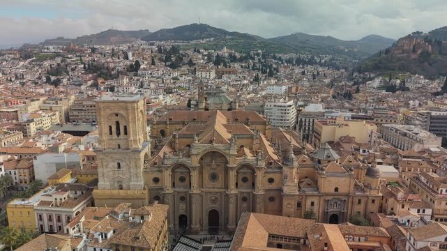 Huge Renaissance Cathedral and Moorish Alhambra complex stand in Granada, Spain. Terracotta roofs fill dense urban cityscape of ancient Andalusian town. 4K aerial of distant Spanish mountains