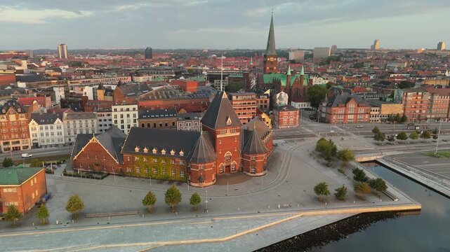 Dense historic red brick architecture and towering green cathedral spire dominate Aarhus, Denmark. Calm sea water reflects traditional coastal harbor in Danish city. 4K aerial of urban Orhus skyline