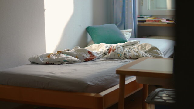 Unmade child bedroom bed with crumpled blanket and pillow near window suggesting morning routine and quiet domestic space with traces of recent sleep and daily life
