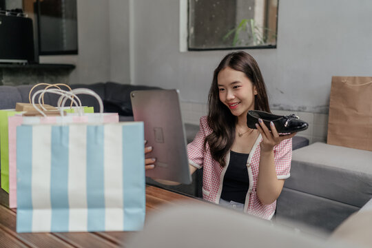 Smiling woman sitting indoors with colorful shopping bags, holding a tablet and displaying a black shoe, suggesting online shopping and fashion choice in a modern living room