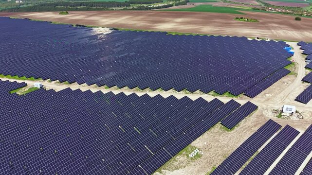 A large area filled with solar panels is seen in a field. The panels are arranged in rows, and the landscape shows patches of green fields and trees.