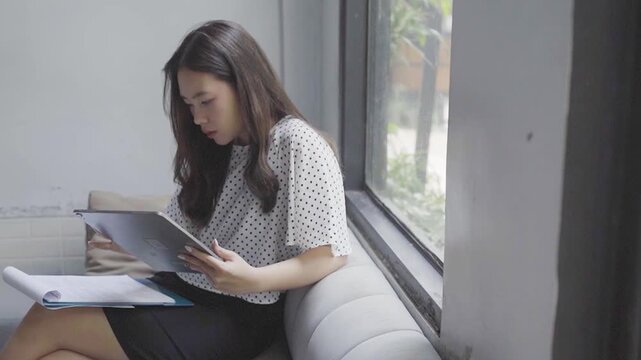 Asian woman proofreading documents with tablet, soft window light, focused expression, annotating pages and marking corrections,
