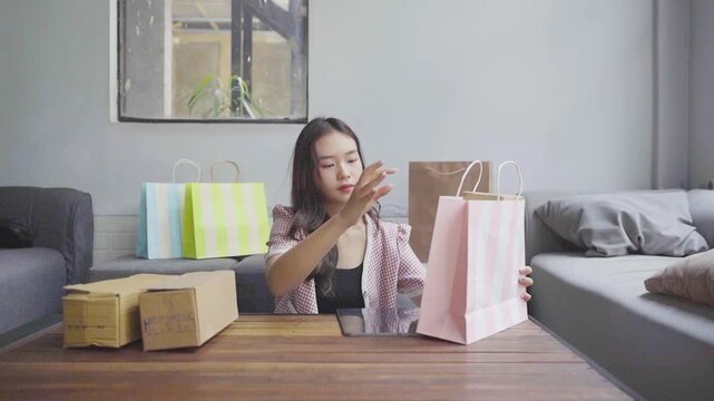 Asian woman packing small business orders on table, cardboard boxes and shopping bags, checking inventory list on phone, focused