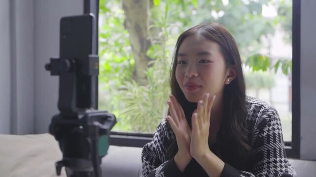 Asian woman storyteller speaking passionately on camera, gesturing with hands, natural window light, phone on tripod, sofa
