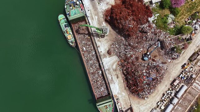 Workers are busy sorting and managing scrap materials at a recycling facility next to a green river. The site shows various types of waste and equipment used for processing.