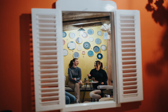 Two friends sit at a small cafe table, talking and smiling while sharing drinks. A casual conversation unfolds against a bright, plate-decorated wall in a warm interior