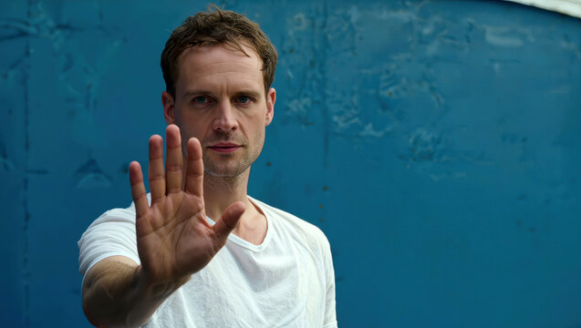 Stop gesture close-up with man raising open palm against blue wall for refusal message, personal boundaries, warning notice and communication training.