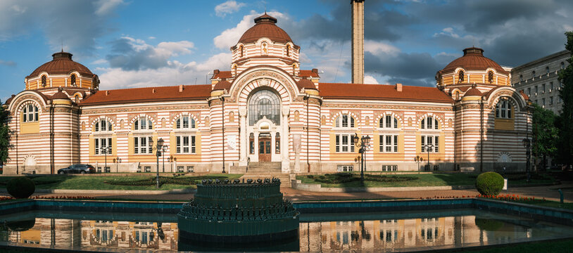 Frontal view of the historic Central Mineral Baths building in Sofia, showcasing its vibrant secessionist facade and stone domes reflected on the fountain pool under a summer sky, Bulgaria