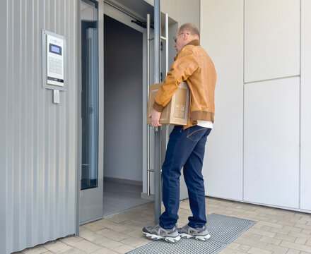 Man holds cardboard box at a modern apartment entrance. He stands by an open glass door and access panel in daylight. Delivery and relocation service concept..