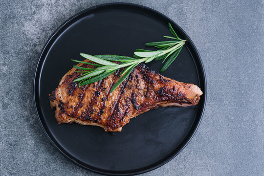Grilled meat served on a black plate with fresh herbs on top on a concrete background. The juicy meat features distinct grill marks and a golden-brown crust. top view