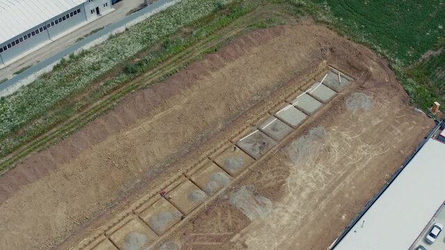 Wide aerial drone shot of rectangular excavation pit with concrete foundation slabs and timber formwork for future commercial building between existing industrial facilities. Perfect for construction 