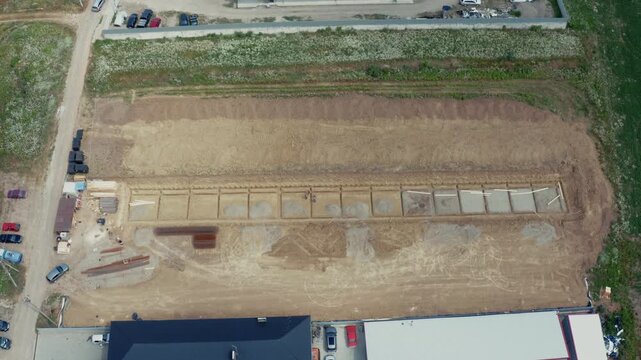 Wide top-down aerial drone shot of large excavation pit with concrete strip foundation and timber formwork for future commercial building, surrounded by construction vehicles and materials. Perfect fo