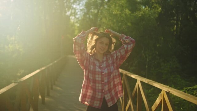 A young woman stands centered on a wooden bridge with her arms outstretched in warm sunlight, surrounded by lush green trees and a peaceful outdoor landscape