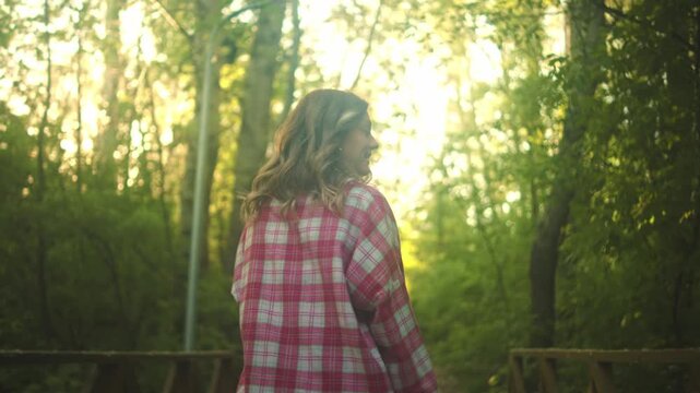 A smiling woman stands on a wooden bridge in a green forest, holding her hands behind her head in soft daylight with warm sunlight filtering through the trees