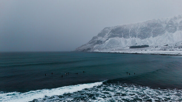 Aerial view of surfers waiting for waves in the dark Atlantic waters near snow-covered mountains under a moody winter sky in Unstad, Nordland, Norway.