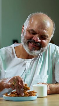 Senior Indian man eating pakoda or pakora with tea on table, Asian foodie old man expressing immense happiness while enjoying tasty onion fritters in modern kitchen setting, joyful retired person