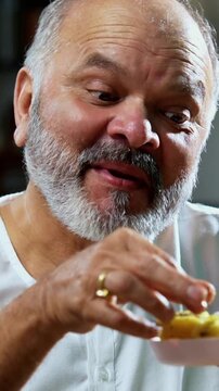 Senior Indian man eating khaman dhokla with joyful expression, Asian foodie old man enjoying tasty Gujarati snack while sitting at table in modern kitchen, expressing immense happiness and delight