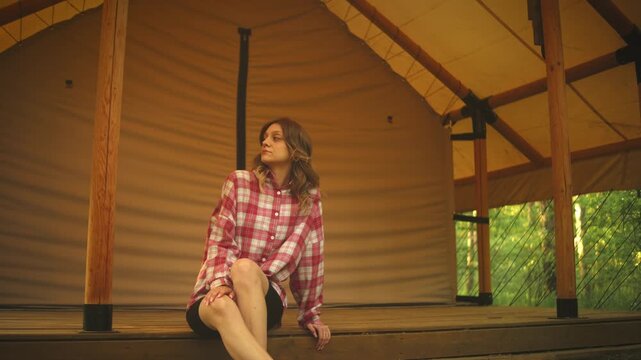 A woman in a plaid shirt sits on a rustic wooden porch beside a canvas shelter, facing the camera in a relaxed pose. Soft sunlight, green foliage