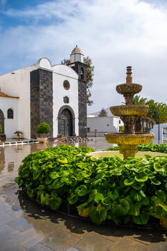 A fountain surrounded by green plants is in front of San Bartolome Church in Lanzarote. The scene shows rain on the ground and cloudy skies above