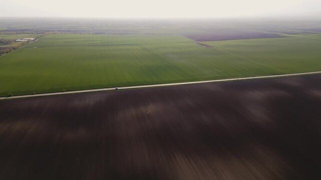 Drone shot flying over a plowed soil and green fields as a white car drives along a rural road. High angle aerial view of agricultural landscape and road trip concept.