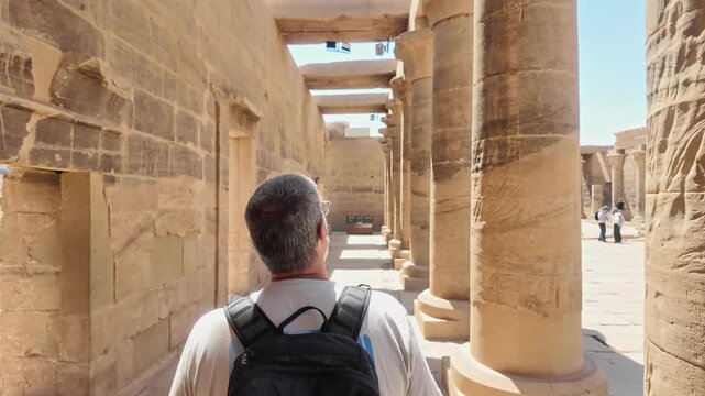Middle aged tourist man with a backpack walking through the colonnade of the ancient philae temple, a famous archaeological site in aswan, egypt
