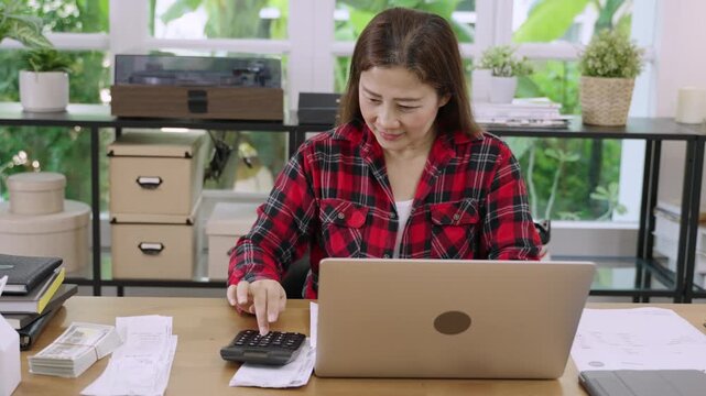 Professional Asian female accountant sat at desk with laptop computer using calculator to calculate company bills including wages utilities and taxes in office. Businesswoman working in her workplace.