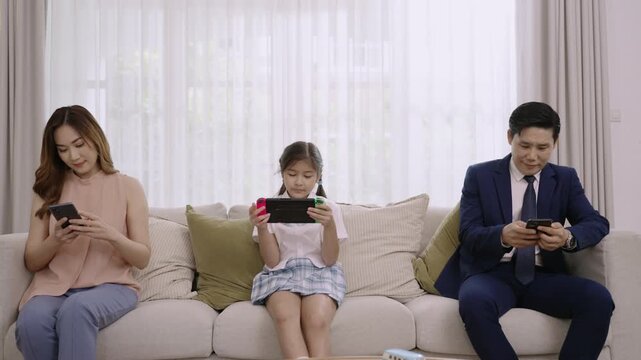 Asian family devices addicted to and overuse social media, so they are not paying attention to each other father and mother playing smartphones while daughter play game console on sofa in living room.