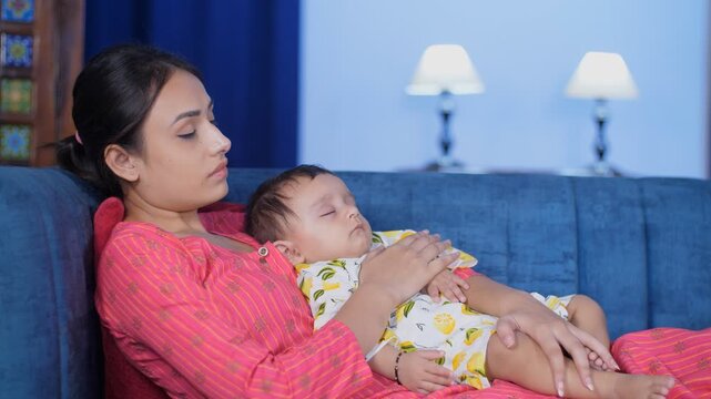 A tired Indian mother and her little baby girl resting together on the sofa. Young mother helping her infant to fall asleep by patting her gently - work flexibility, role model, parenting, tiring sche