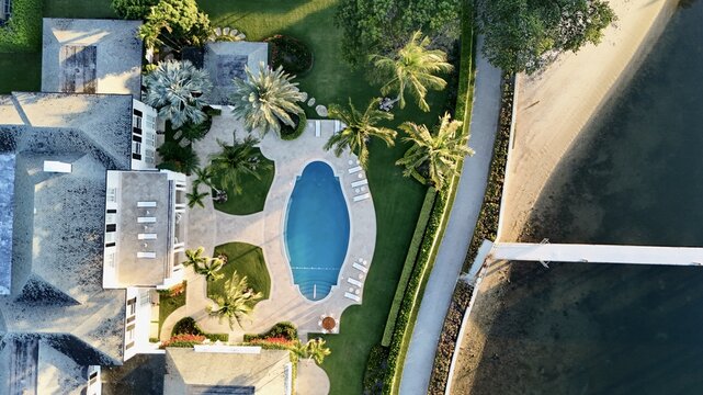 Aerial view of a luxury waterfront estate featuring a blue swimming pool, lush palm trees, and a private dock along the coast in Palm Beach, Florida, United States.