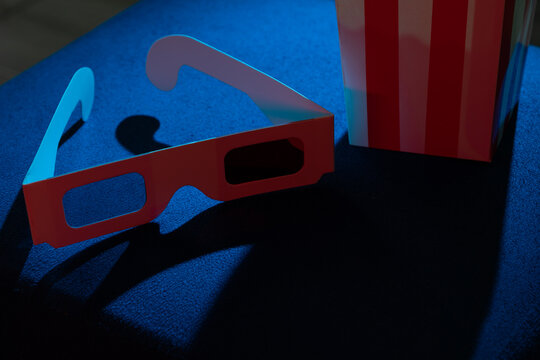 3D glasses next to a box of popcorn on a chair in a dark setting. Concept of movies, entertainment, leisure, and fun moments at home