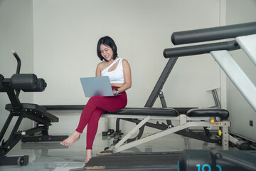 Fototapeta premium Smiling young Asian woman in sportswear sitting on a fitness bench and using a laptop, working remotely, checking her workout plan, or managing an online fitness business in a gym