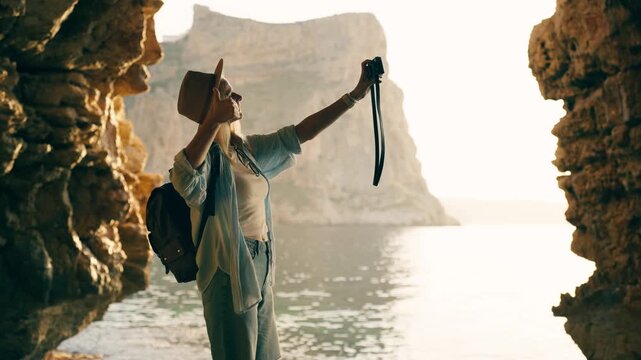 Adventurous female traveler exploring sea cave and taking selfie with vintage camera, enjoying ocean sunset during vacation, representing wanderlust, travel lifestyle, and summer adventure.