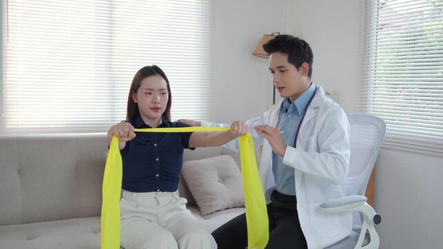 Young Asian male doctor assisting a female patient with physiotherapy exercises using an elastic band for shoulder and arm rehabilitation