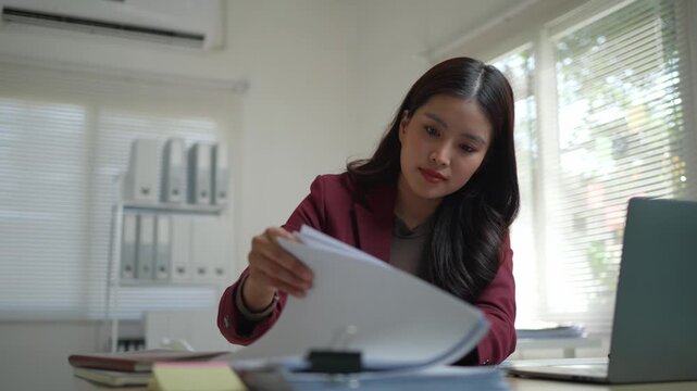 Professional asian woman sitting at her desk, diligently working on her laptop and sorting through important company paperwork