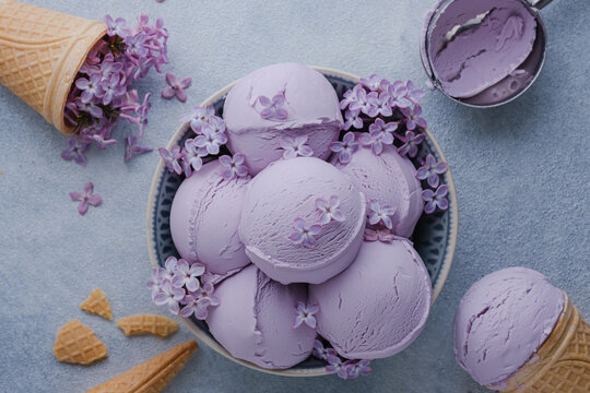 A bowl holds scoops of lavender ice cream decorated with lilac flowers on a blue background. Healthy frozen dessert. top view