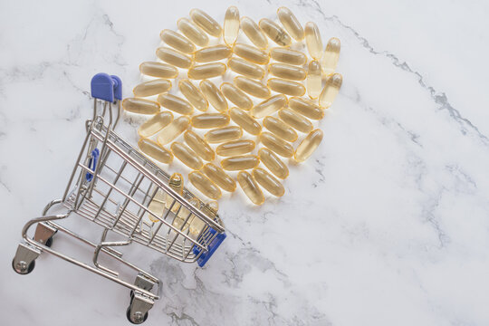 High angle view of a small shopping cart overturned with golden fish oil gel capsules scattered on a white marble surface, conceptualizing medicine purchase