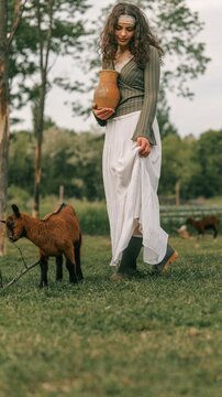 Young woman in a long skirt holding a clay jug walks with a goat in a grassy field