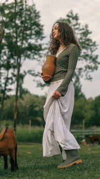 Young woman in rustic attire holding a clay jug in a grassy field with goats