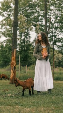 Young woman in a white skirt and green top holding a jug in a field with a goat
