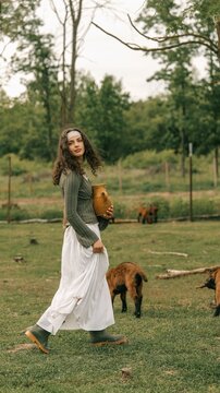 Young woman in traditional dress carrying a clay jug walks through a field with goats