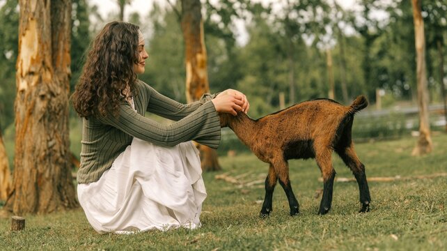 Young woman gently petting a small brown goat in a grassy field