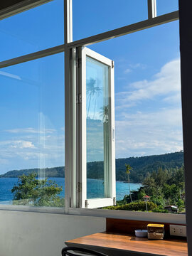 POV from inside a room looking through an open white window to a stunning tropical ocean view with palm trees and hills